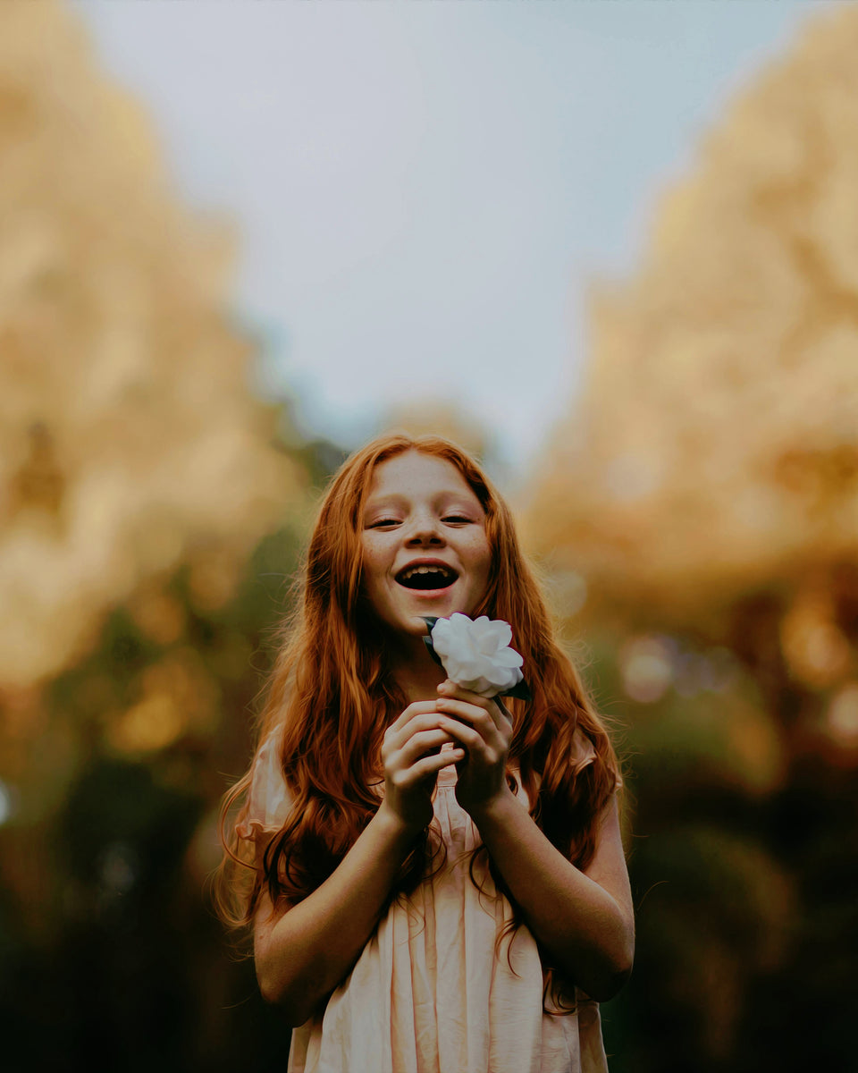 Young girl with red hair holding a white flower in a blurred outdoor setting